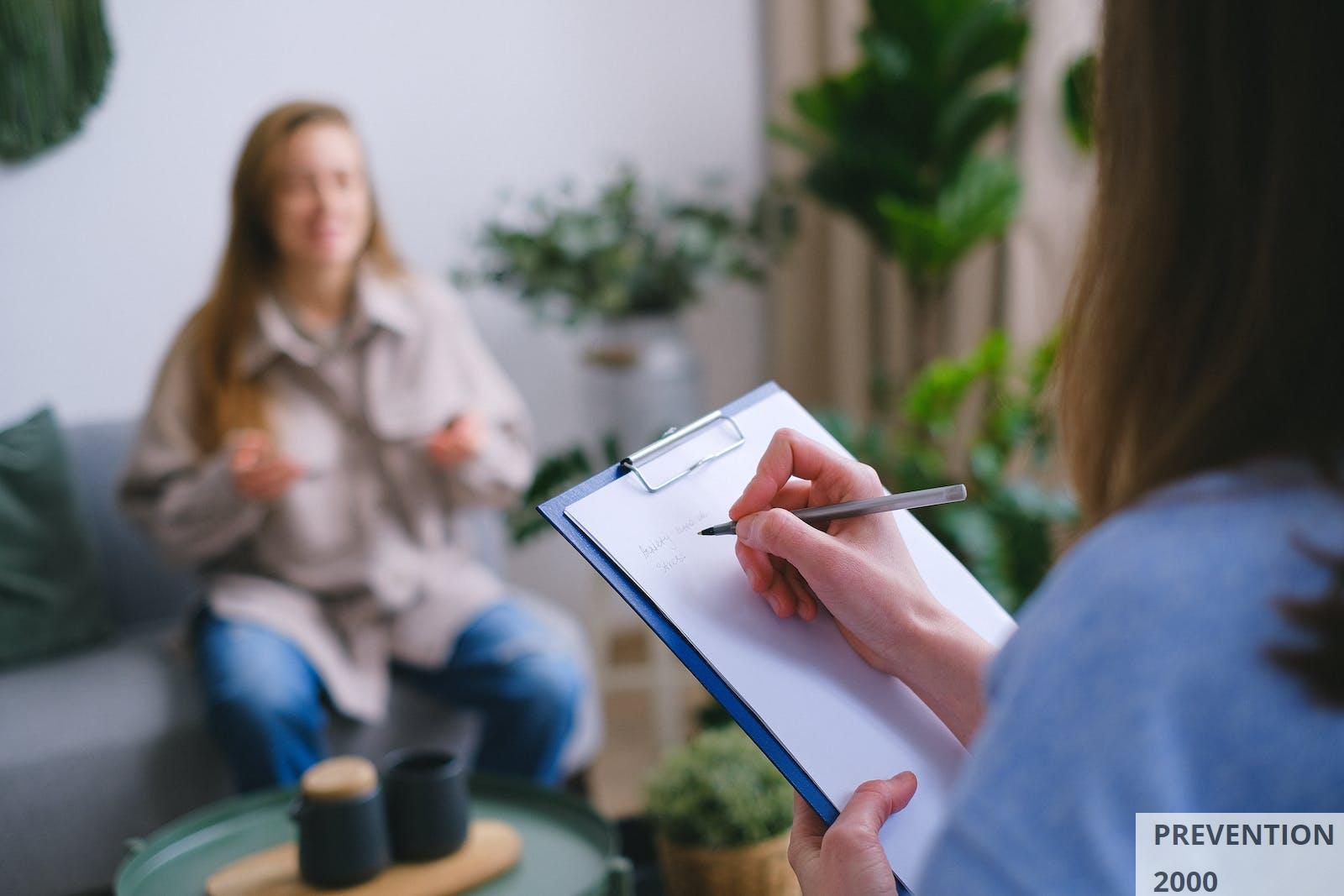 Unrecognizable professional female psychologist writing on clipboard while sitting against client on blurred background during psychotherapy session in light office
